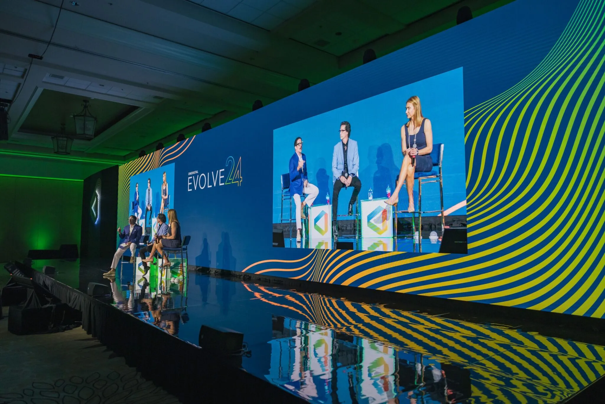 OneDigital 'Evolve 24' event stage with three panelists seated on stools in front of a large blue and yellow LED backdrop.